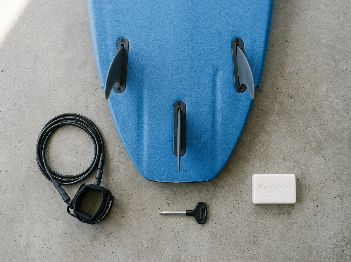 Foam soft top surfboard tail with three soft fins, a fin key, coiled leash and a bar of surf wax on concrete